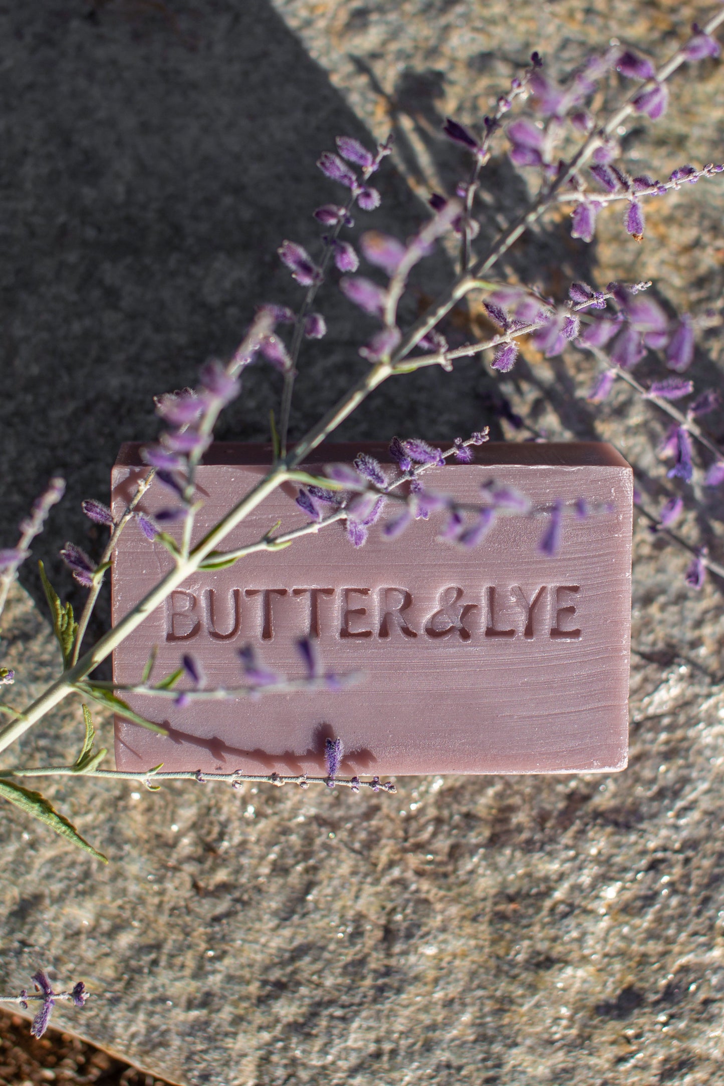 A rectangular purple bar of soap with the brand name ‘Butter & Lye' engraved on its surface. The soap rests on a textured stone background, and a sprig of purple flowers partially drapes across the soap, casting shadows.