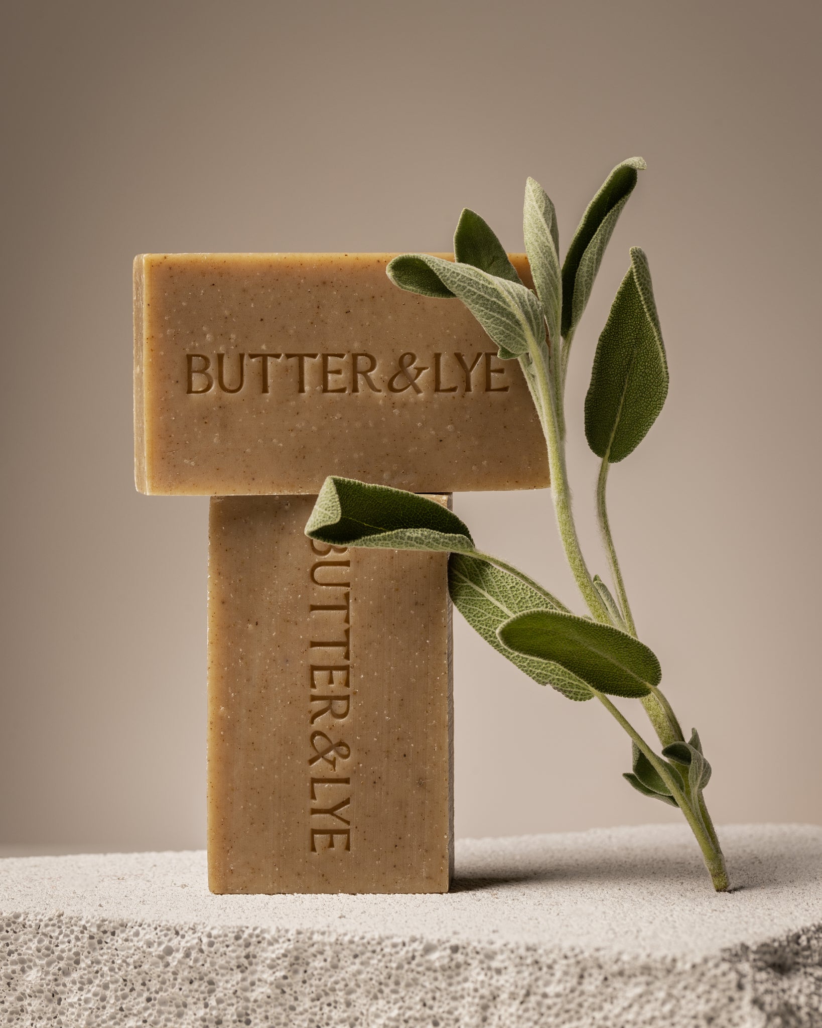 Two rectangular bars of sage soap stacked on top of each other in the shape of the letter “T”, with a sprig of fresh sage leaves resting between them. The bars are a light golden brown with "Butter & Lye" embossed in capital letters.