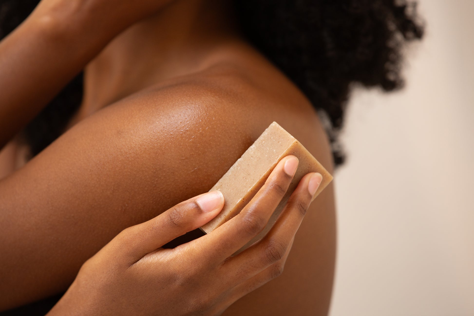 A close-up shot of a person's hand and shoulder, showing a light brown rectangular soap bar with "Butter & Lye" embossed on it being held against their skin. The background is a soft, blurred neutral tone.