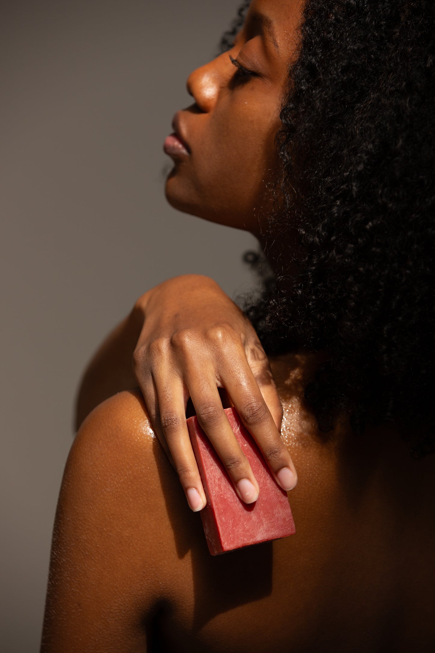 A close-up of a person’s shoulder and back, where the person is holding a red bar of soap and pressing it gently against their wet skin. The soft lighting accentuates the smooth texture of the skin and the soap.