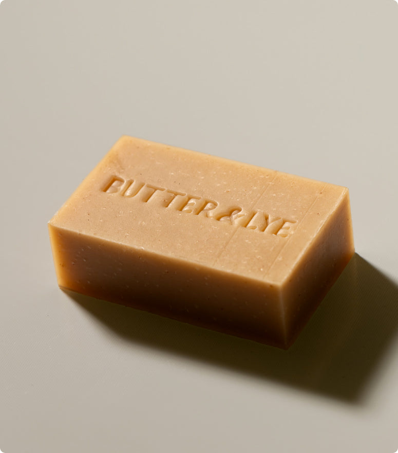 A studio-style shot of a light brown rectangular soap bar, labeled "Butter & Lye," resting on a clean, neutral-colored surface. The image is taken from a slightly elevated angle.