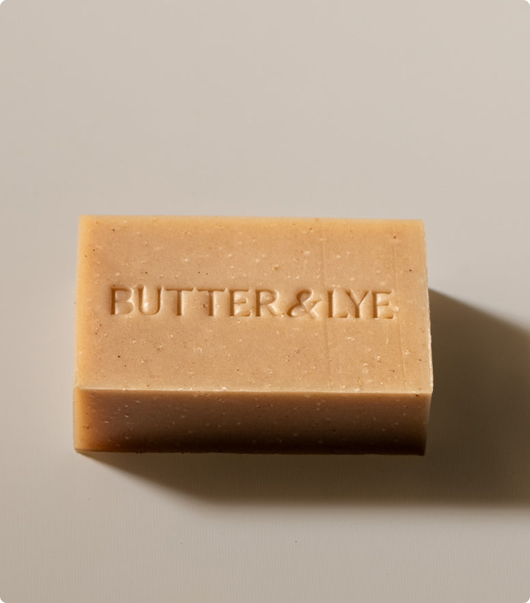 A close-up, top-down shot of a rectangular bar of soap, light golden brown with a slightly darker base. The words "Butter & Lye" are embossed in capital letters on the top surface.