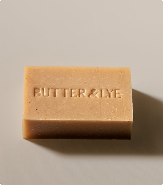 A close-up, top-down shot of a rectangular bar of soap, light golden brown with a slightly darker base. The words "Butter & Lye" are embossed in capital letters on the top surface.