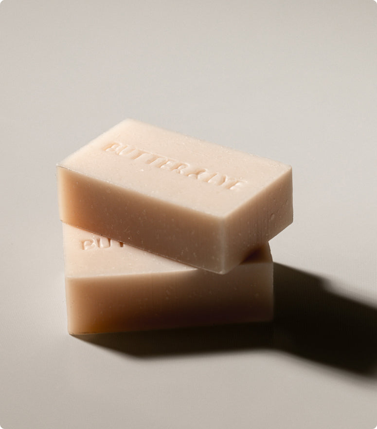 Two rectangular bars of light-colored, creamy soap are stacked on top of each other against a light background. The top bar has the word Butter & Lye embossed on its surface.