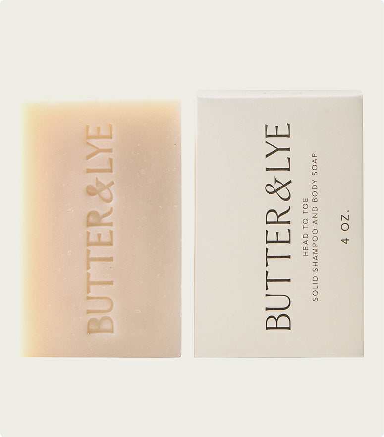 A close-up shot of a rectangular bar of all in one soap alongside its packaging. The soap is packaged in a simple, brown paper wrapper with black lettering that reads “Butter & Lye - Head to Toe solid shampoo and body soap 4 oz”.