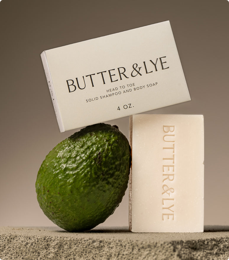 Butter & Lye soap bar displayed alongside its minimalist packaging and a fresh green avocado. The all in one soap bar, embossed with the brand's name, is suitable as a face and body soap, offering a natural and gentle cleansing experience.
