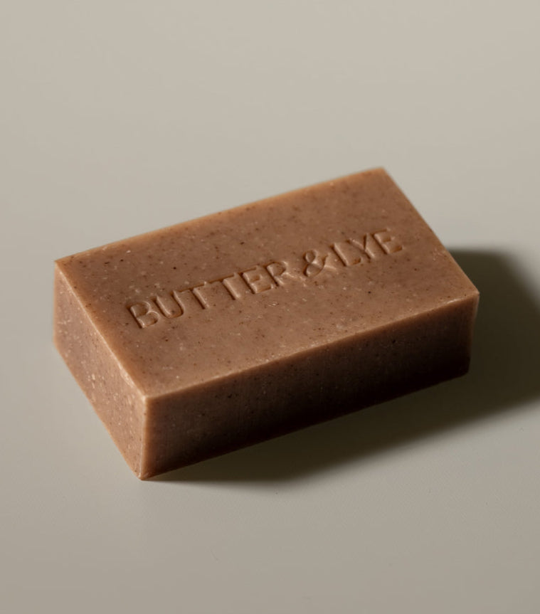 A close-up of a rectangular brown Butter & Lye soap bar with a smooth texture, placed on a neutral background.