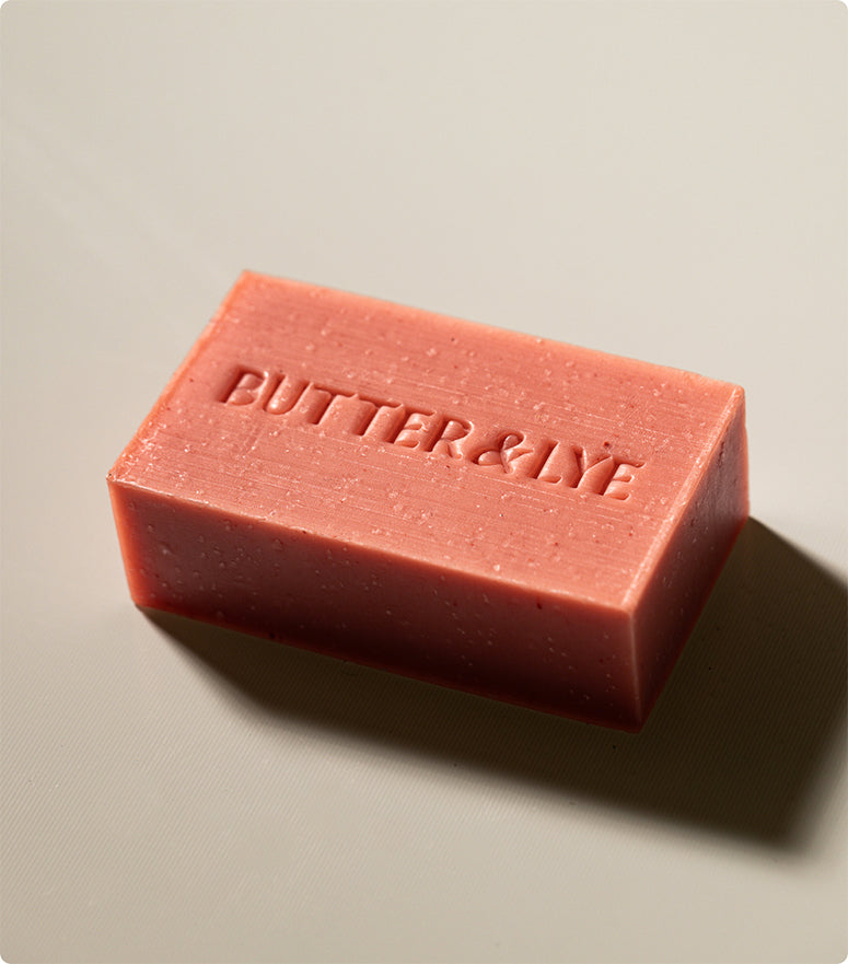 A single pink soap bar embossed with "Butter & Lye," resting on a neutral beige surface, showcasing its natural, handcrafted texture.