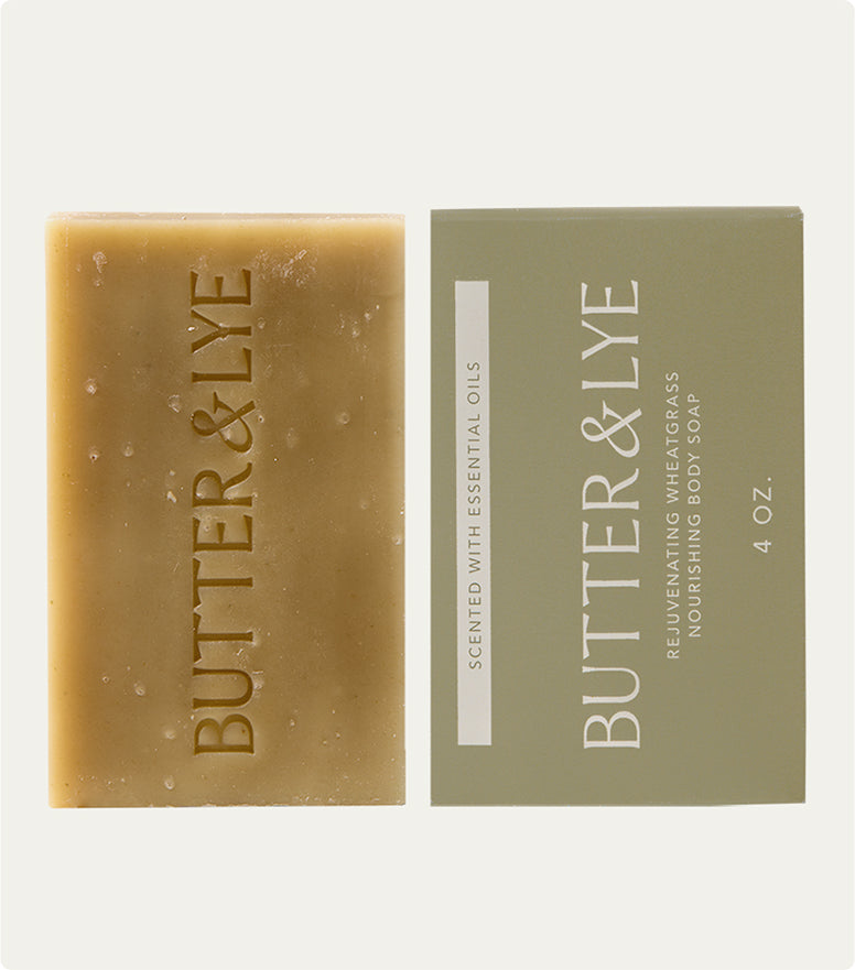 A Butter & Lye wheatgrass soap bar alongside its minimalist packaging. The soap is greenish-brown with a natural texture, while the packaging highlights its nourishing and essential oil-infused formula.