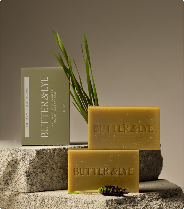Aesthetic display of Butter & Lye soap bars on textured stone with a natural backdrop. Two greenish-brown soap bars are arranged alongside a matching packaged box labeled "Rejuvenating Wheatgrass Nourishing Body Soap." A sprig of lavender and fresh wheatgrass leaves enhance the earthy, organic theme.