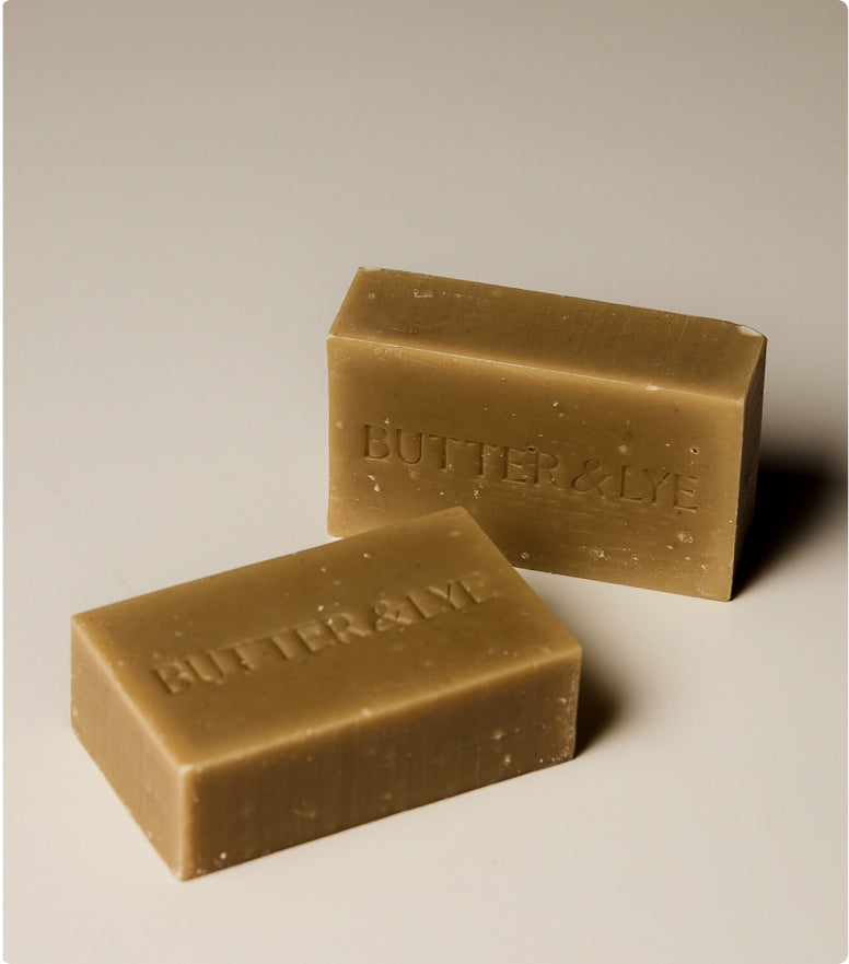 Two greenish-brown Butter & Lye soap bars with a natural texture, embossed with the brand name, placed on a neutral background with soft lighting.