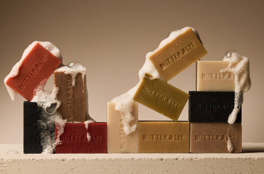 Assortment of Butter & Lye soap bars stacked in a playful arrangement, with light foam and bubbles on some soap bars. Showcasing a variety of natural soap including the best soap for eczema and colors including black, red, green, and beige