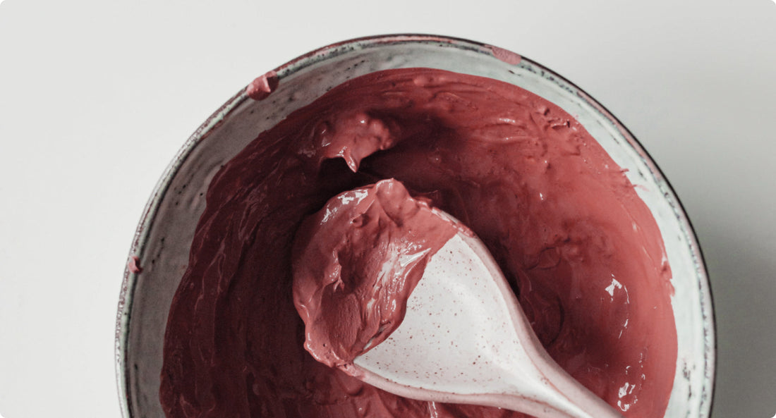 A close-up of a bowl filled with smooth, rich pink clay, with a white spoon scooping up a portion. The clay has a creamy texture and a deep rosy hue, contrasting with the neutral background.