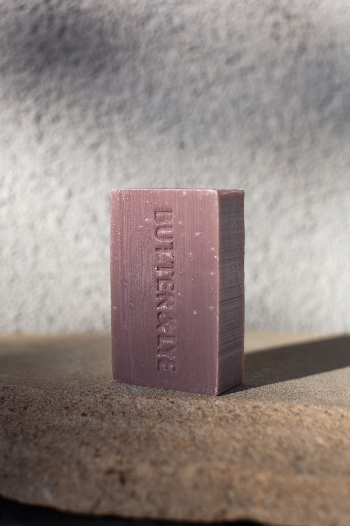 A rectangular purple clay soap bar with a deep, rich purple hue. The words ‘Butter & Lye' are embossed on the front in a slightly lighter shade of purple. The soap is resting on a textured concrete surface.