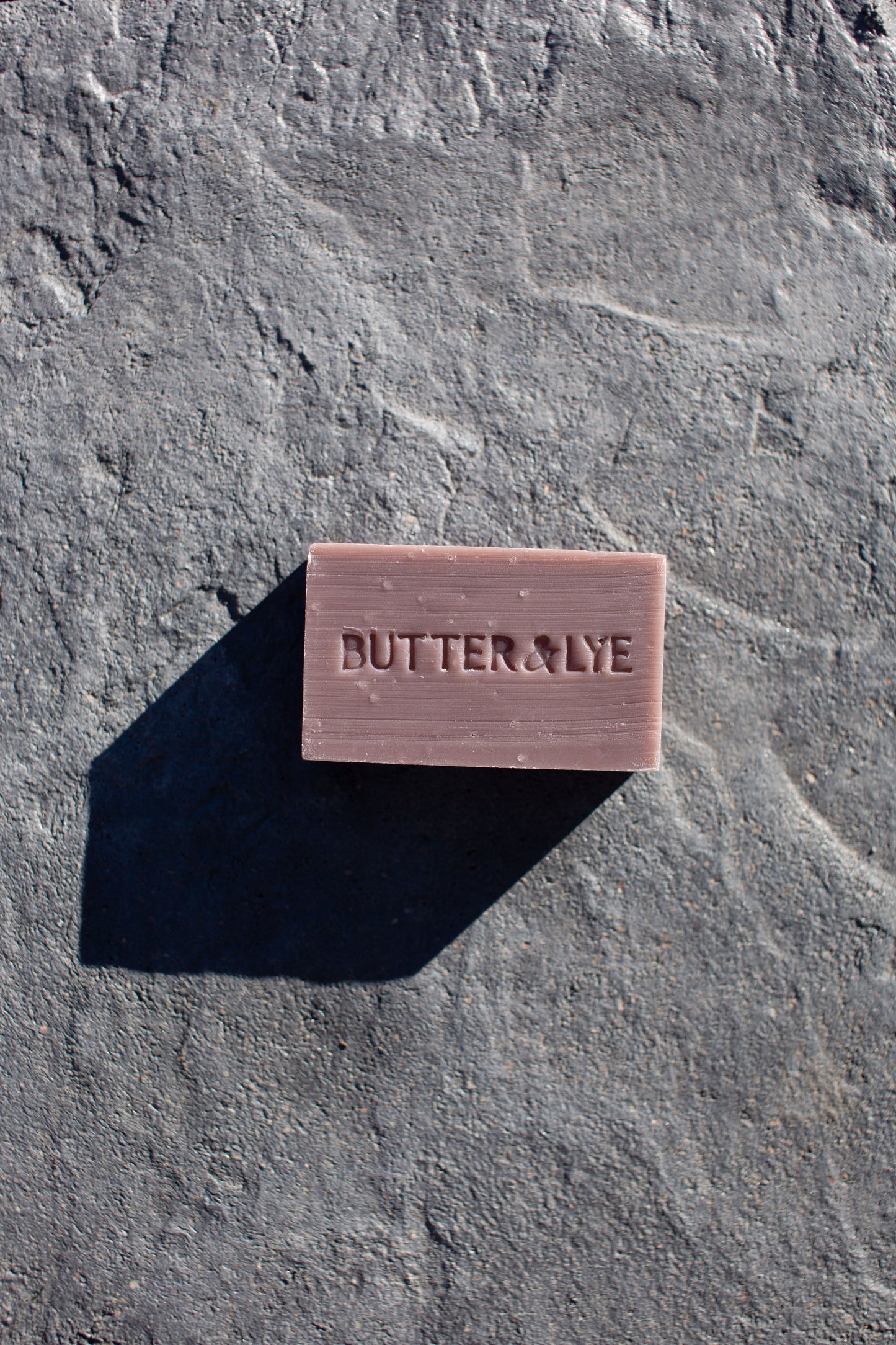 A rectangular purple soap bar with ‘Butter & Lye' engraved on its surface is placed on a textured gray stone background. The soap casts a sharp, long shadow to the left, indicating a light source from the right.