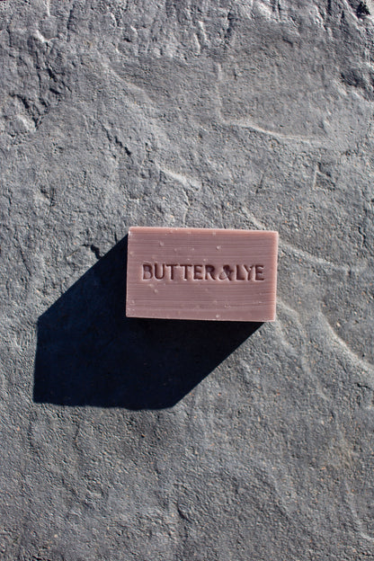 A rectangular purple soap bar with ‘Butter & Lye' engraved on its surface is placed on a textured gray stone background. The soap casts a sharp, long shadow to the left, indicating a light source from the right.