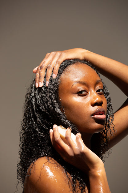 A woman with curly hair looking into the camera with glowing skin, lathering a light cream colored soap bar on her hair and face.