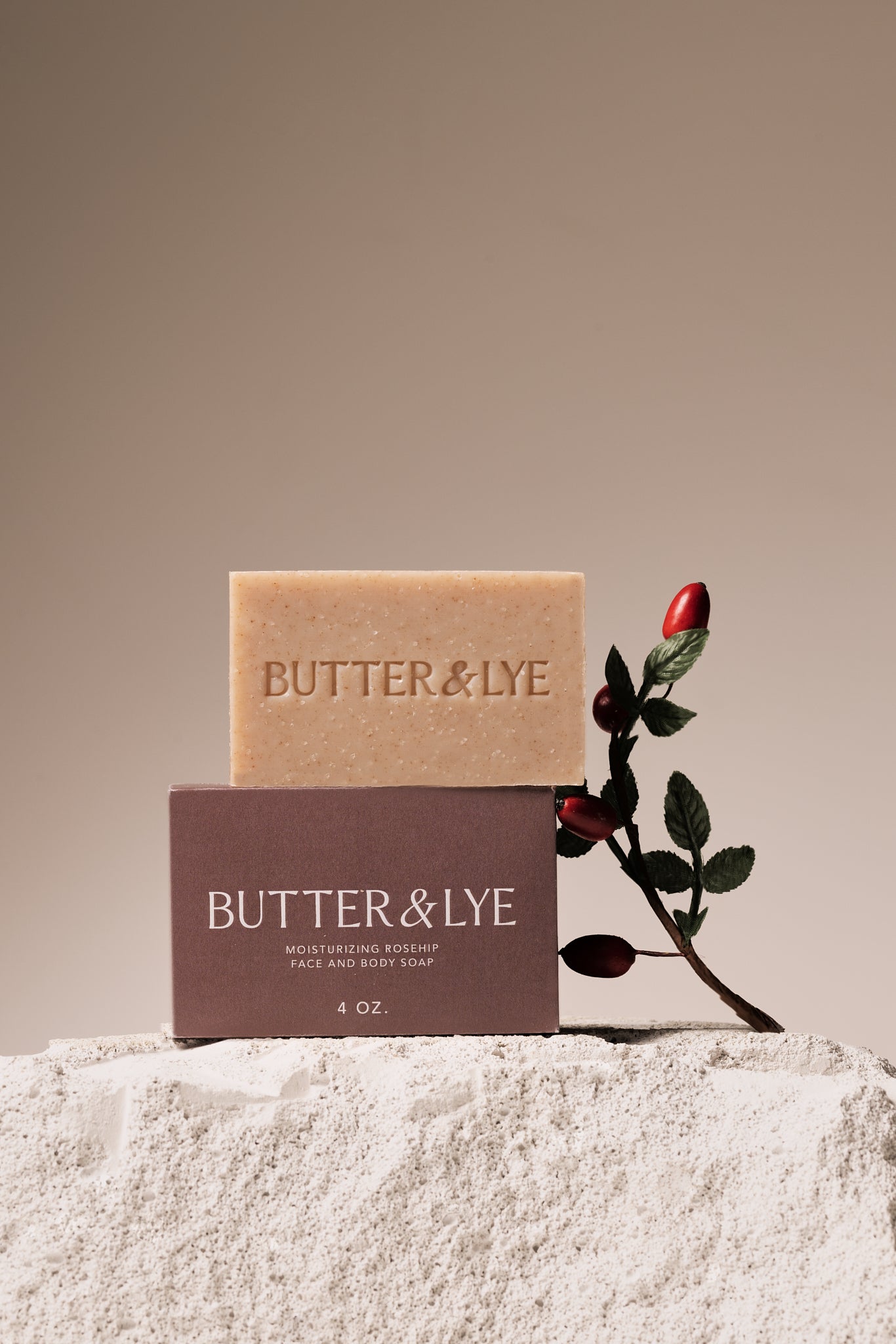 A Butter & Lye rosehip soap bar standing on a rocky surface next to its packaging, with a rosehip branch accenting the natural and luxurious presentation.