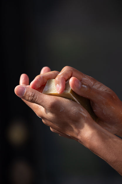 A close-up of hands lathering a Butter & Lye wheatgrass soap bar, showing its rich, foamy texture as it is being used.