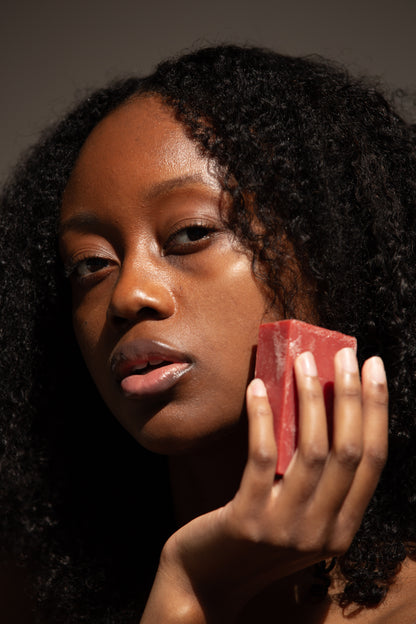 A close-up portrait of a person with dark, curly hair holding a red bar of soap close to their face. The person has a calm, confident expression with soft lighting highlighting their skin and hair texture.