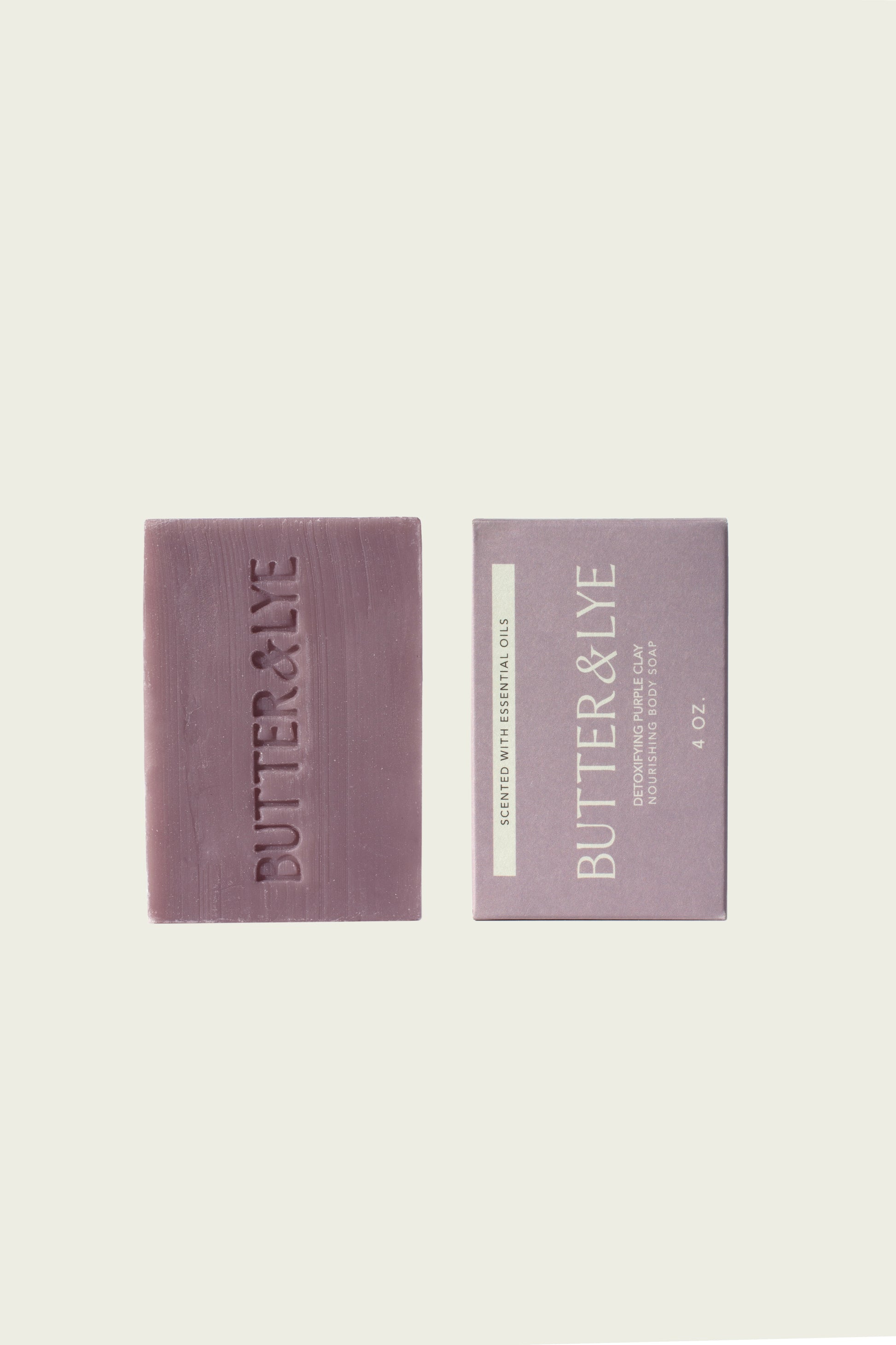 Two rectangular purple soap bars are displayed side by side. The soap on the left has ‘Butter & Lye' engraved on its surface, while the soap on the right features packaging with the same brand name and additional details, including a label with the weight 4 oz and a description of the soap's scent.