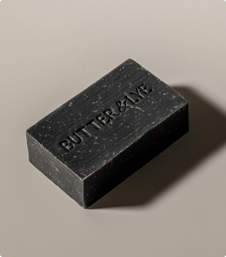 A close-up image of a rectangular black soap bar with "Butter & Lye" embossed on its surface, placed on a light beige background.