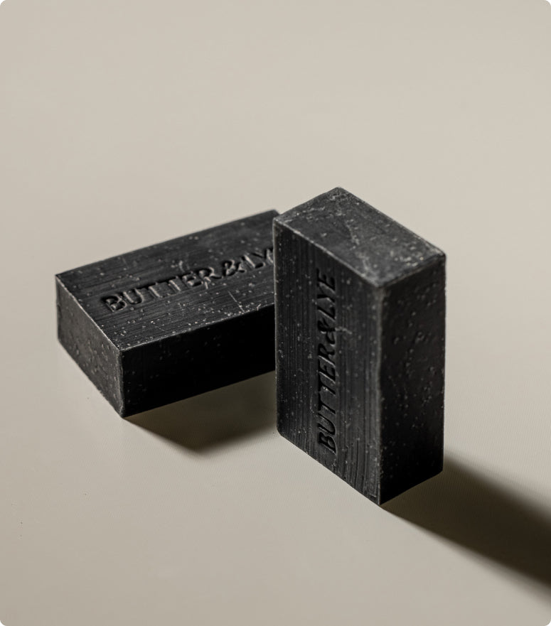 Two black soap bars positioned on a smooth, light surface, casting soft shadows, both featuring the "Butter & Lye" branding.