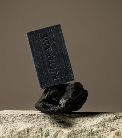 A black soap bar standing vertically on a dark charcoal rock, positioned on a sandy surface with a neutral background, highlighting the "Butter & Lye" branding.