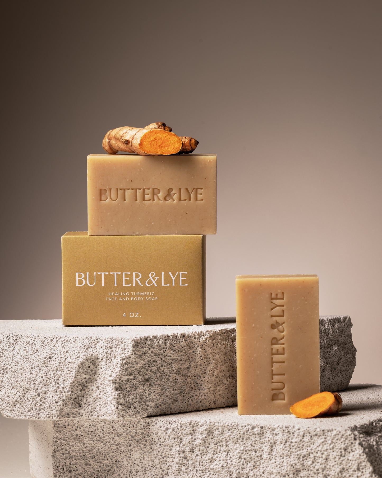 Two bars of turmeric soap with 'Butter & Lye' engraved, resting on textured stone blocks, accompanied by a fresh turmeric root.