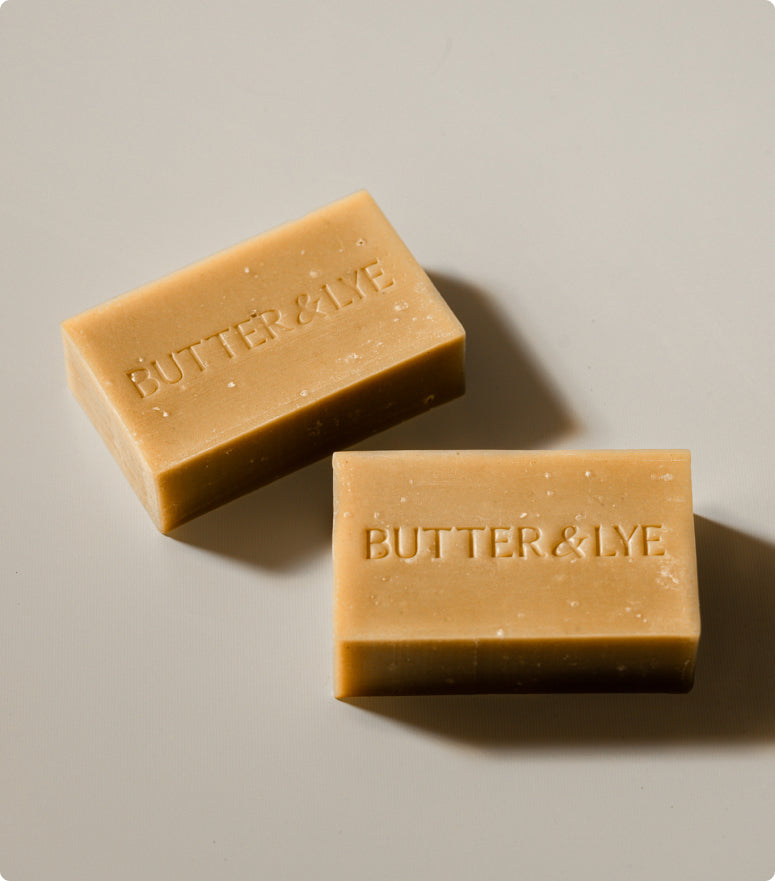 Two rectangular bars of soap with the brand name "Butter & Lye" embossed on them, displayed on a neutral background. The turmeric soap bar has a natural, beige color and a slightly textured surface.