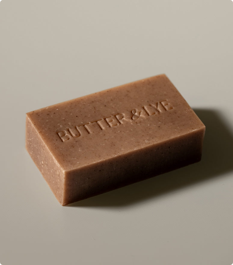 A close-up of a rectangular brown Butter & Lye soap bar with a smooth texture, placed on a neutral background.