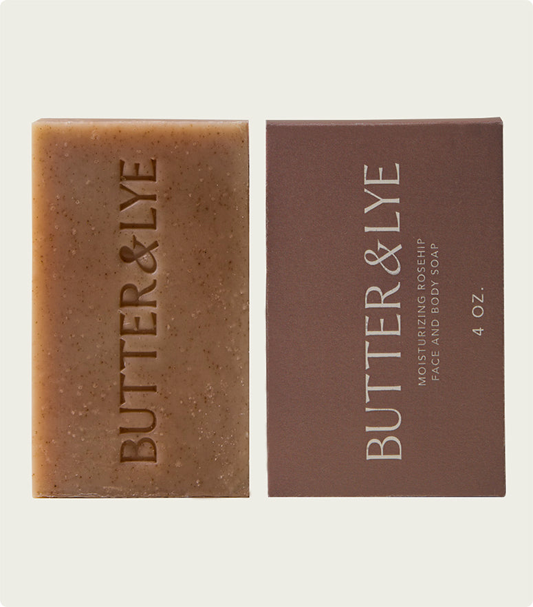 A vertical shot of the Butter & Lye soap bar placed beside its minimalist brown packaging that reads "Moisturizing Rosehip Face and Body Soap, 4 oz."