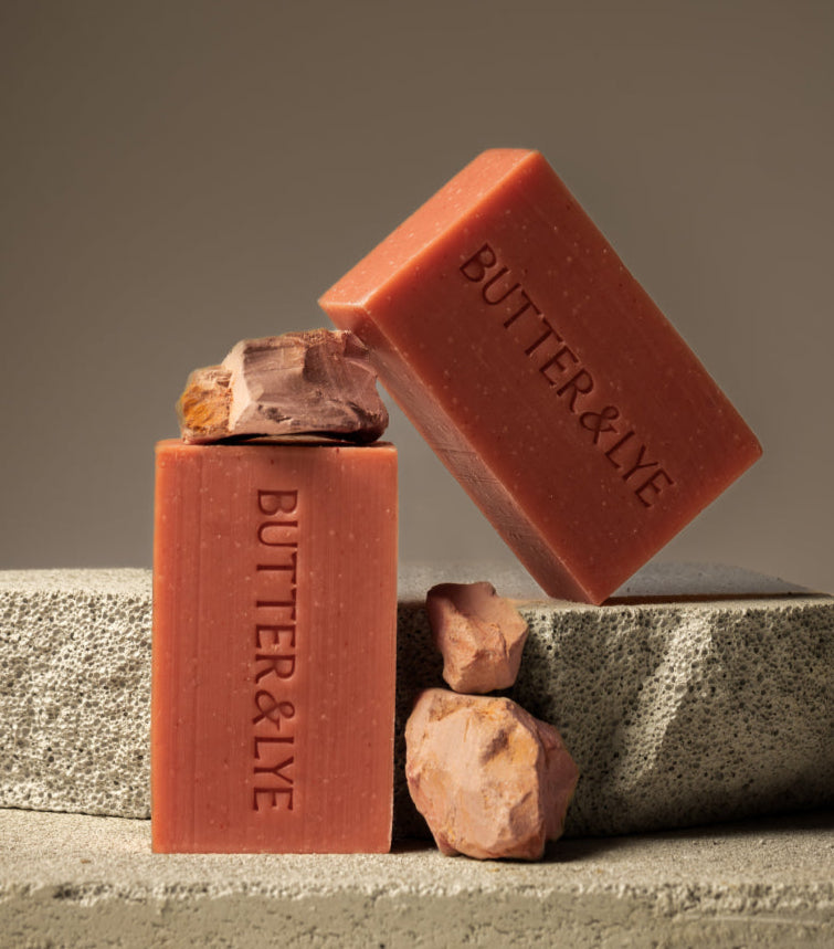 Two pink "Butter & Lye" soap bars arranged on stone blocks, complemented by natural clay rocks, highlighting the soap's earthy and clay-inspired origins.