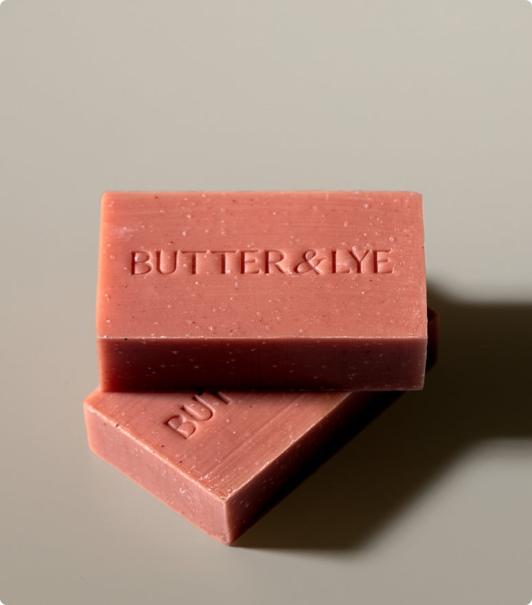 Two bars of "Butter & Lye" reddish-pink toned soap bars stacked on top of each other on a beige background, emphasizing the minimalist design and rich color of the soap.