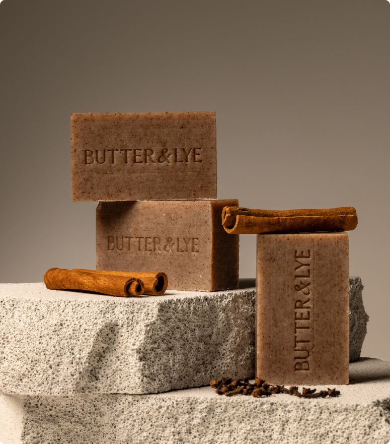 Three bars of the same 'Butter & Lye' embossed soap stacked on rough, stone-like blocks. The scene includes cinnamon sticks and cloves, complementing the soap's earthy, cinnamon-like aesthetic.