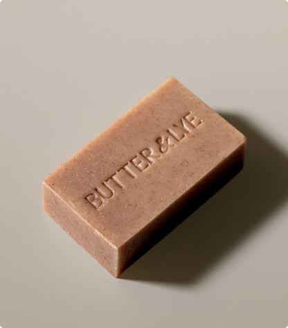 A single bar of brown-colored soap embossed with the words  'Butter & Lye'  resting on a smooth, beige surface. The soap has a natural, handmade appearance, evoking a cinnamon-scented feel.