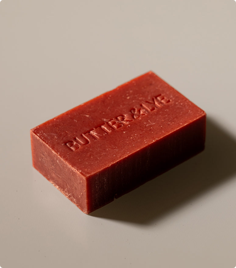 A rectangular, maroon bar of soap with the words "Butter & Lye" embossed on its surface. The soap rests on a neutral beige background, and soft lighting casts a gentle shadow beneath it.