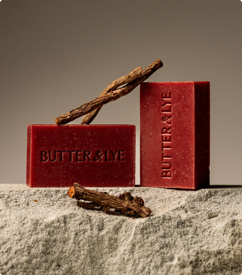 Two red Butter & Lye soap bars displayed on a textured stone surface, with dried plant roots artfully arranged beside and on top of the soap.