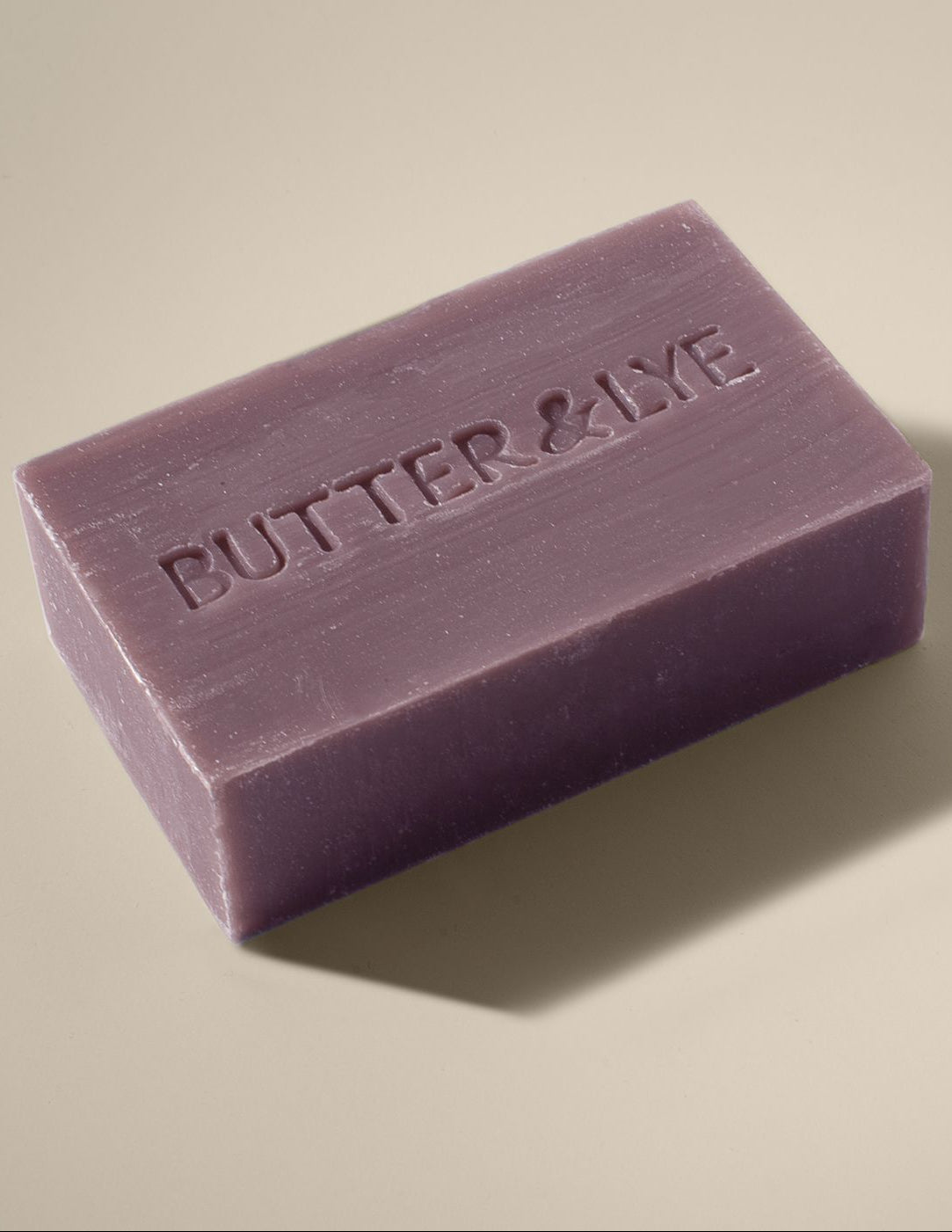 A rectangular, purple clay bar of soap with the words ‘Butter & Lye' embossed on its surface. The soap rests on a neutral beige background, and soft lighting casts a gentle shadow beneath it.