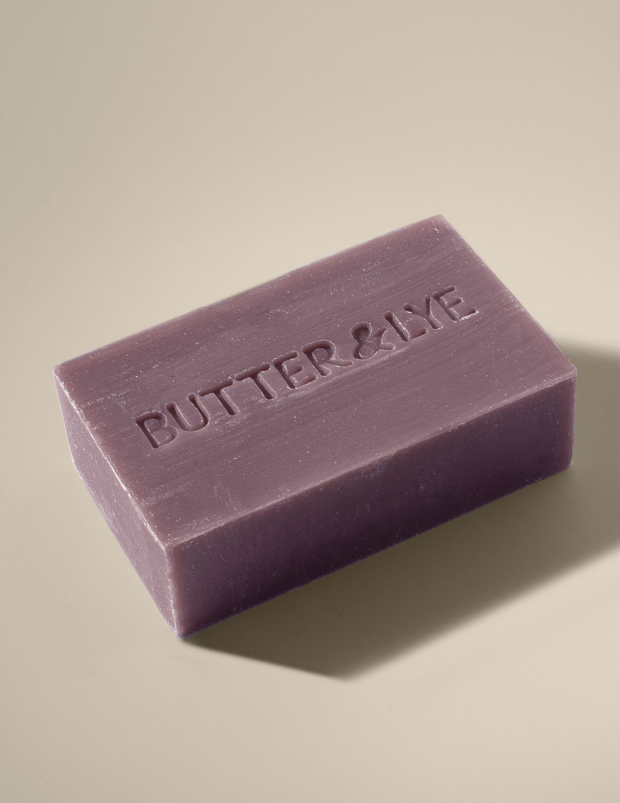 A rectangular, purple clay bar of soap with the words ‘Butter & Lye' embossed on its surface. The soap rests on a neutral beige background, and soft lighting casts a gentle shadow beneath it.